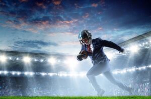 A business man wearing a football helmet and carrying a football runs down a brightly-lit football field.